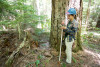 Emily Dewald-Wang holds a PVC pipe which is the target for the laser survey tool (total station). She calls out the species, size, and condition. The data are recorded into a tablet and then the tree's location is mapped with the total station laser.